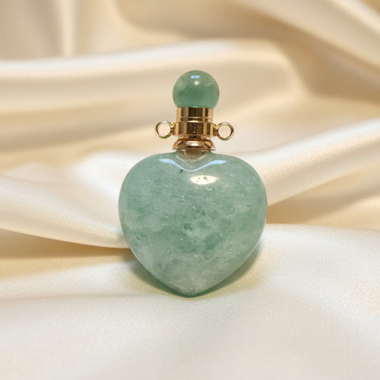 A heart-shaped amazonite  green crystal pendant with a gold-toned cap and green bead, resting on a textured off-white surface.