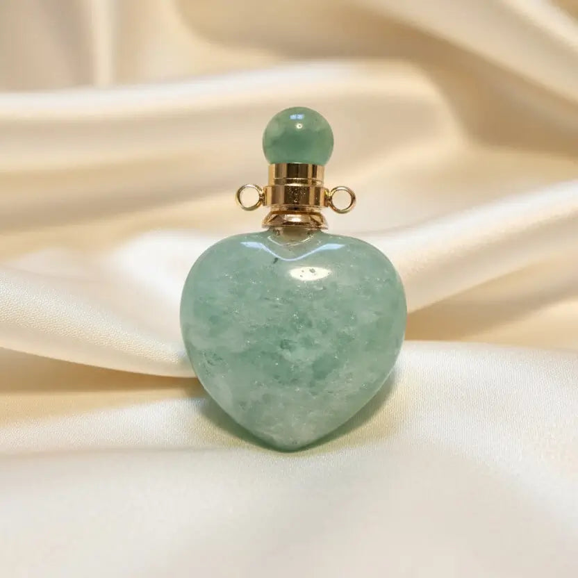 A heart-shaped amazonite  green crystal pendant with a gold-toned cap and green bead, resting on a textured off-white surface.