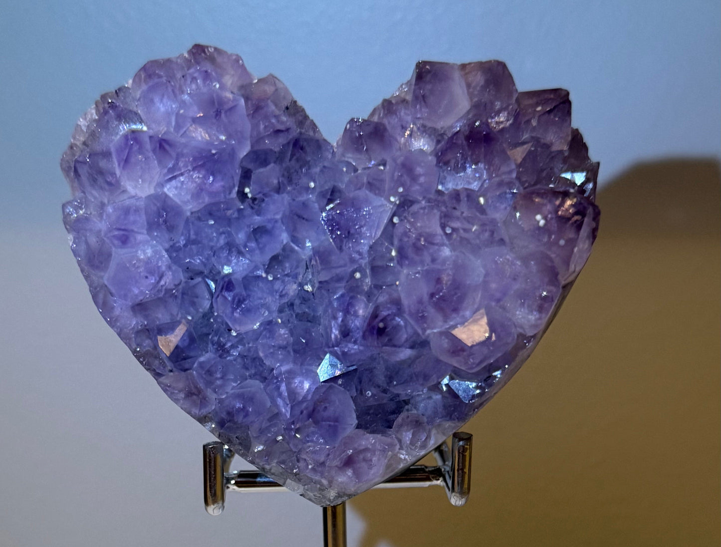 A heart-shaped purple amethyst crystal cluster displayed on a clear acrylic stand, resting on a white 
circular display 