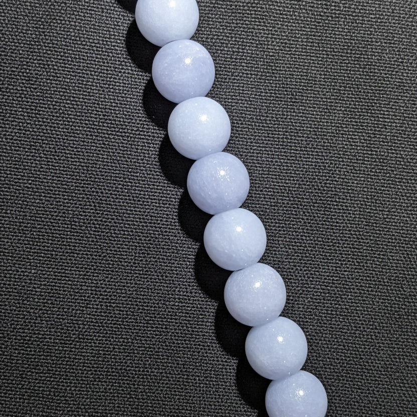 A close-up of a strand of smooth, Angelite blue-gray round beads, likely arranged vertically on a black surface