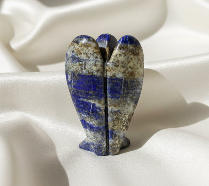 A polished lapis lazuli stone object with a heart-like or shell-like form, featuring deep blue bands over a beige and brown marbled base, photographed on a light off-white surface.