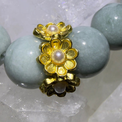 A close-up of a beaded aventurine bracelet featuring smooth, pale blue-green round beads and a central gold-tone floral charm with a pearl-like center, displayed on white crystal formations.