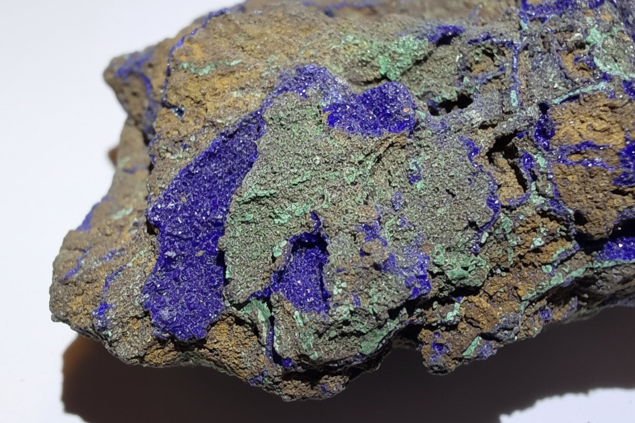 A close-up of an azurite mineral specimen with layered purple, green, and brown crystalline textures on a white background.