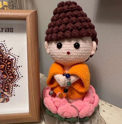 Crochet Buddha doll in orange robes, seated on a pink lotus flower, holding a chakra bead chain.