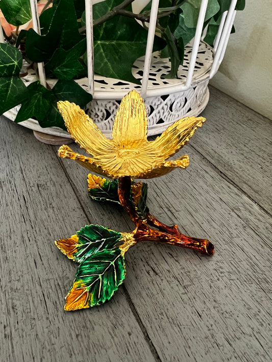Decorative golden flower with green and gold leaves on a wooden surface. White decorative cage with greenery in the background.
