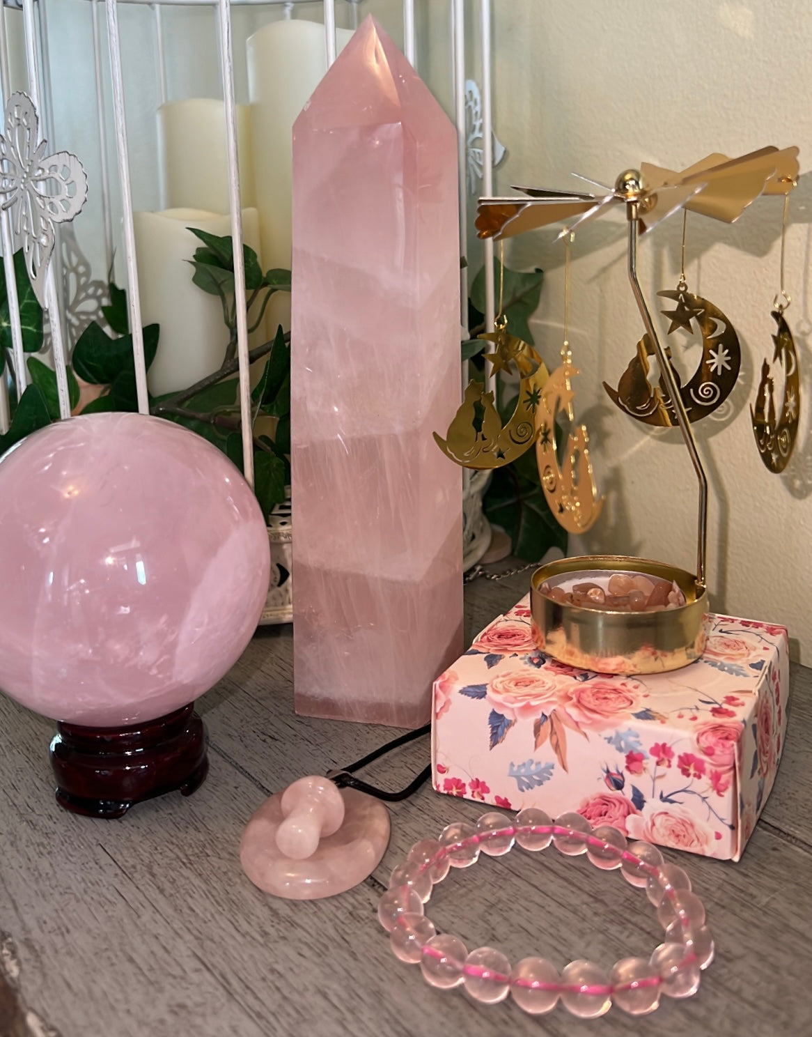 Still life of various pink rose quartz crystals including a tower, sphere on a stand, a bracelet, and a donut-shaped charm; a floral box with a gold candle holder and hanging gold moon and cat charms.