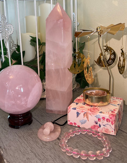 Still life of various pink rose quartz crystals including a tower, sphere on a stand, a bracelet, and a donut-shaped charm; a floral box with a gold candle holder and hanging gold moon and cat charms.