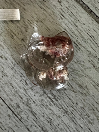 Crystal cat figurine with red and white inclusions on a gray wood surface.