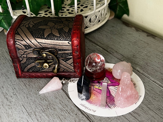 Overhead shot of a wooden treasure chest next to a plate of crystals. There is a clear crystal ball, black crystal, rose quartz Buddha statue, and several other smaller pink crystals. 