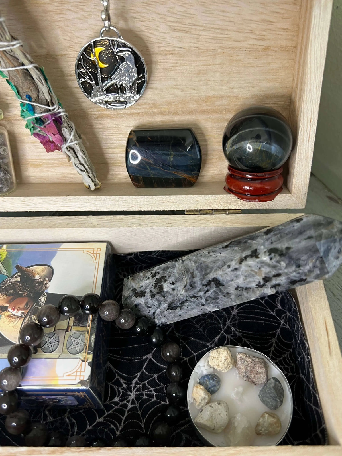 Wooden box filled with crystals, sage, tarot cards, and a crow pendant. 
