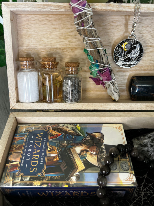 Wooden box containing salt, oil, crystals, sage bundle, raven necklace, tiger's eye, Wizards Tarot deck, and obsidian bracelet.

