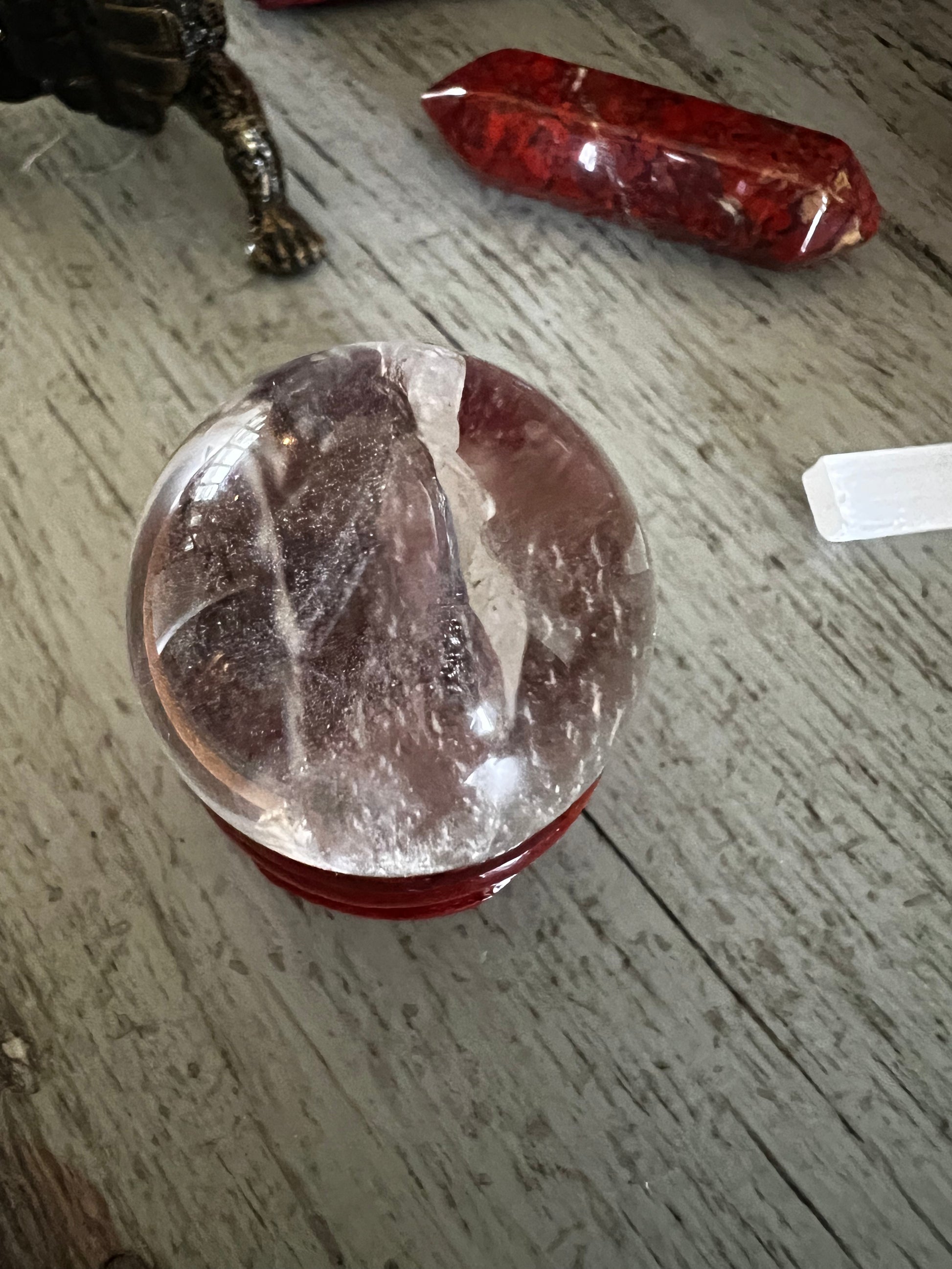 A crystal ball rests on a red stand, with a red crystal and a white selenite wand nearby on a wooden surface.