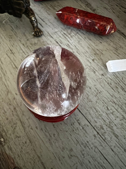 A crystal ball rests on a red stand, with a red crystal and a white selenite wand nearby on a wooden surface.