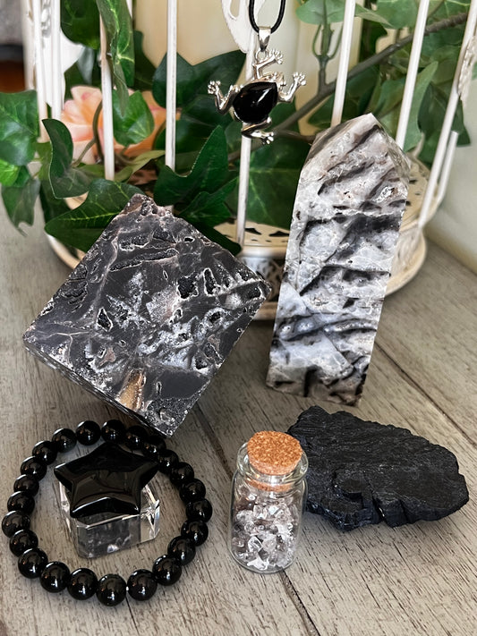 Natural Crystals and jewelry arranged on a wooden surface. A necklace with a silver frog pendant, a gray and black square crystal, a tall gray and white crystal point, a black beaded bracelet with a star-shaped gem, a small glass jar with a cork, and a piece of black crystal are displayed. A white decorative birdcage with greenery and a pink flower is in the background.

