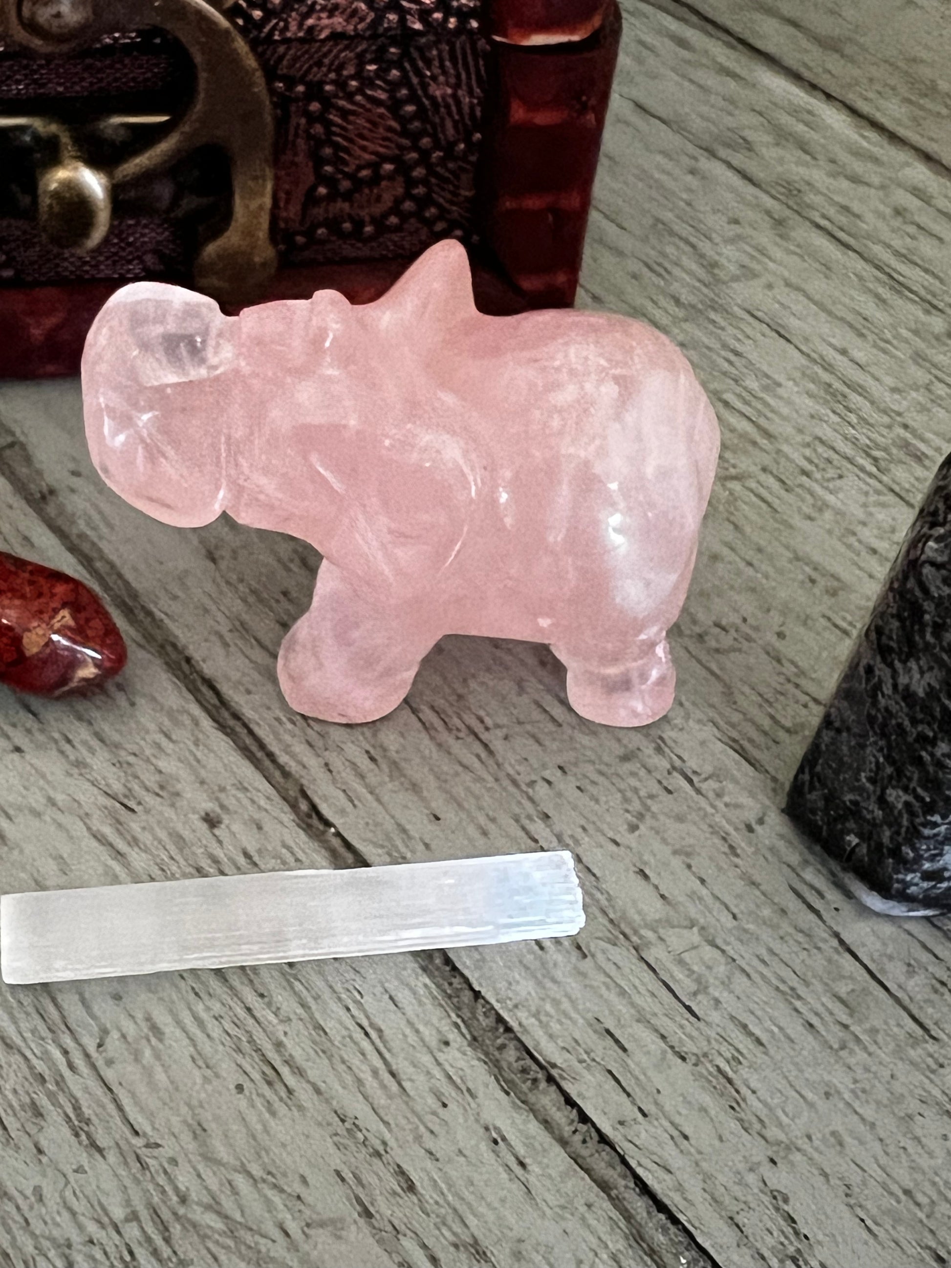 Close-up of a pink crystal elephant, a red stone, a white crystal bar, and a black stone on a gray wooden surface.