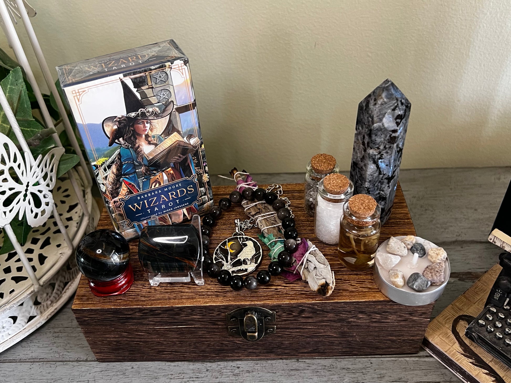 Still life featuring the Barbara Moore Wizards Tarot deck, crystals, sage, and vials arranged on a wooden box with a metal latch. The tarot deck depicts a woman in a hat and blue jacket reading a book. Other items include a black crystal sphere on a red base, a tower crystal, and a beaded necklace with a raven pendant. A miniature typewriter sits to the right.