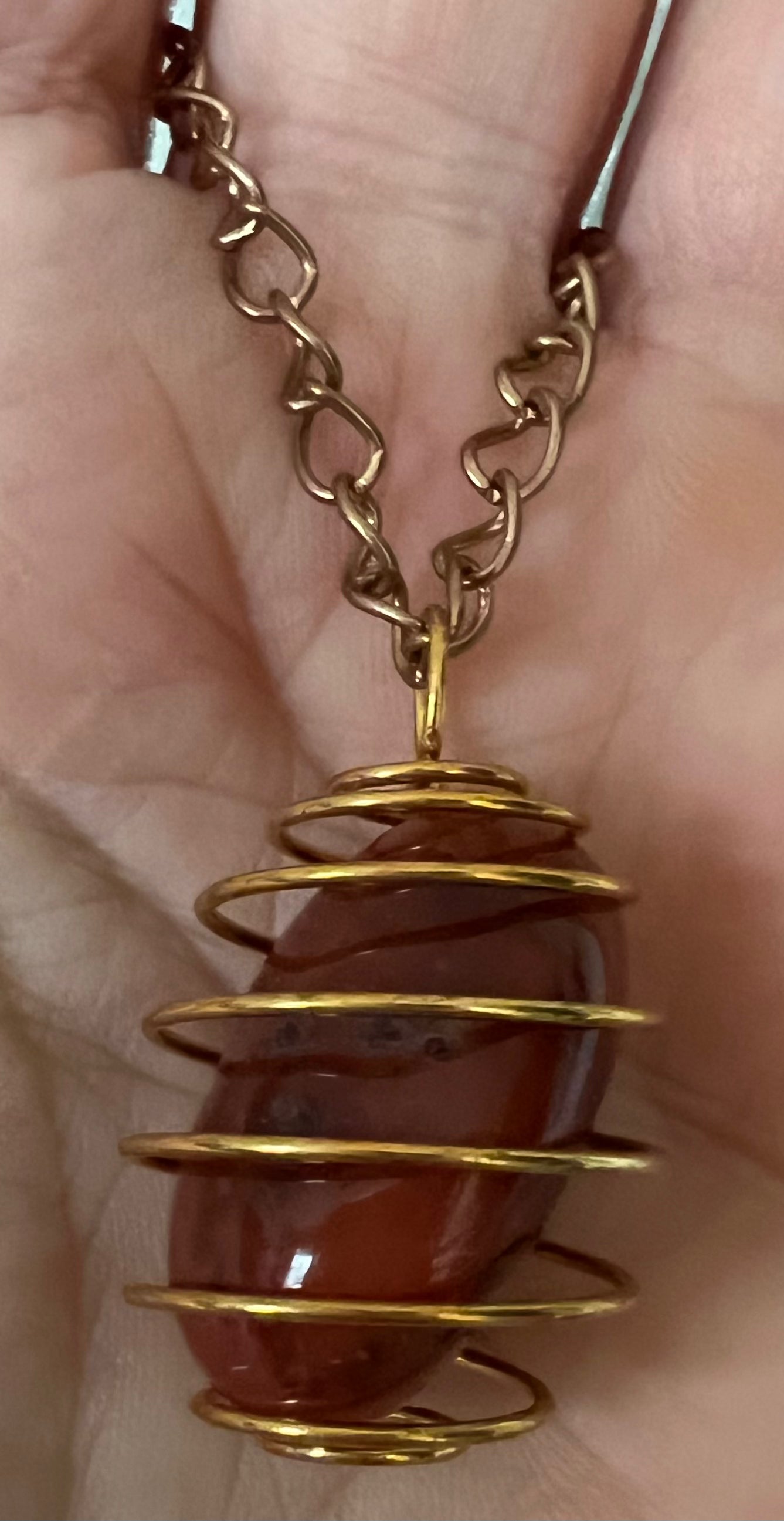 Close-up of a carnelian gemstone pendant wrapped in gold wire on a gold chain, held in a person's palm.
