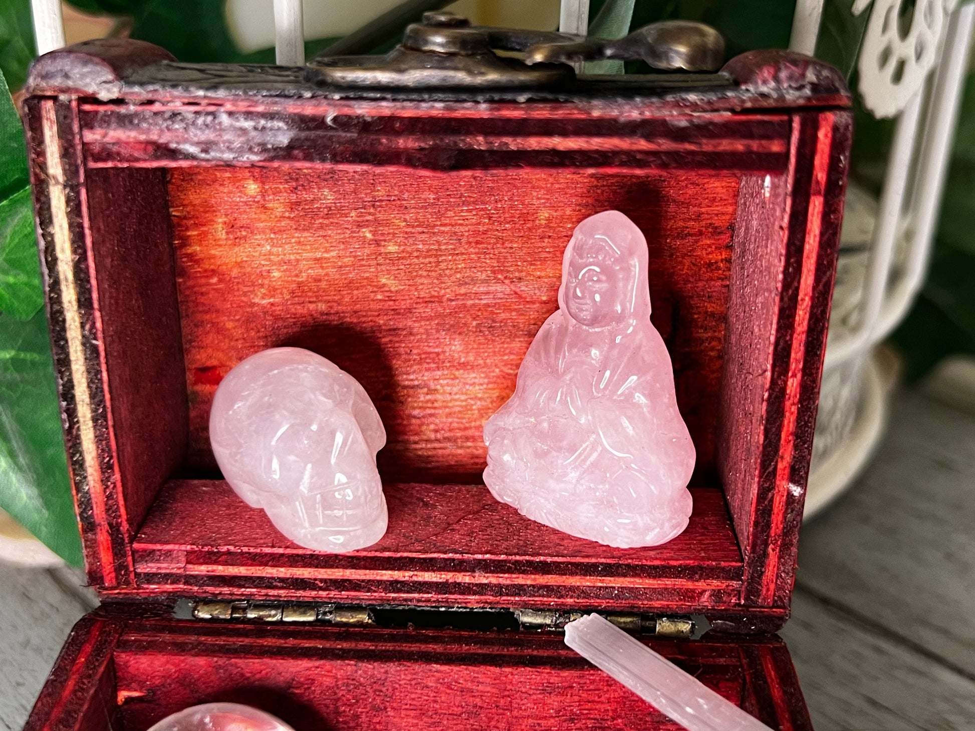 Close-up of a small, open, red wooden box containing two rose quartz figurines, one skull and one Buddha, along with a crystal wand in the box's lower compartment.