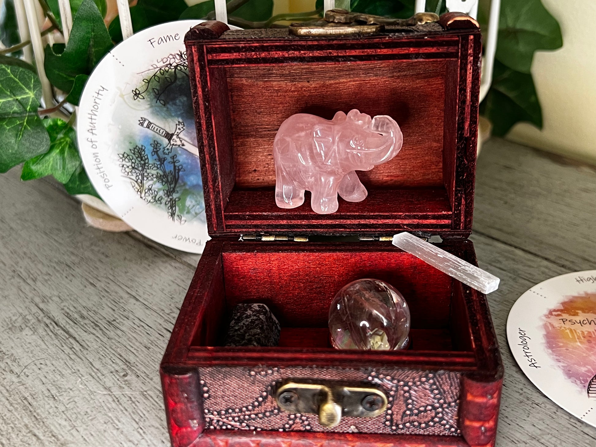 An open, decorative wooden box filled with a rose quartz elephant carving, a selenite wand, and two stones, sitting next to oracle cards and greenery on a wooden surface.
