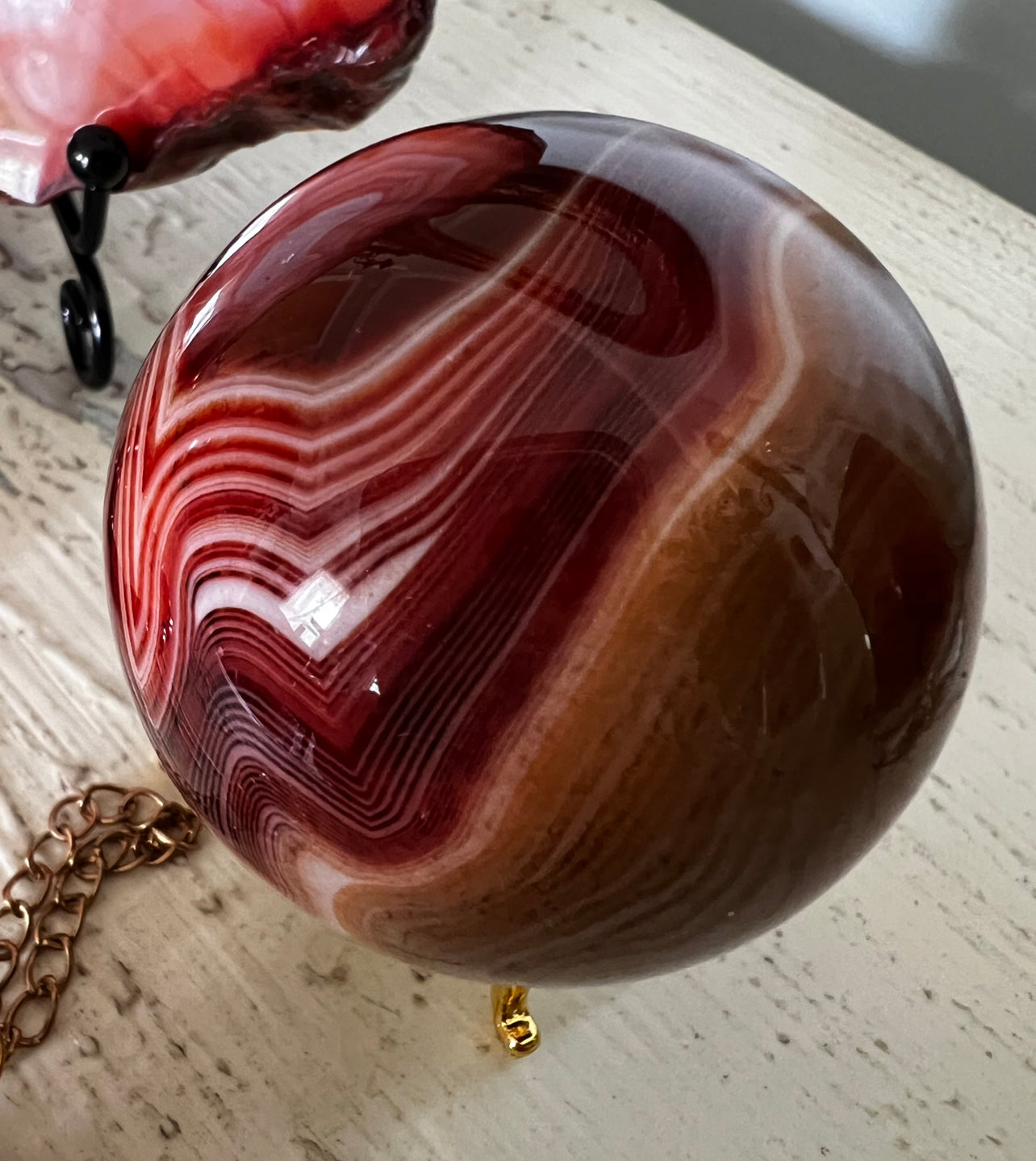 A polished red and brown agate sphere with white stripes, resting on a gold tripod stand with a gold chain nearby.