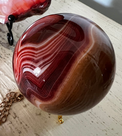 A polished red and brown agate sphere with white stripes, resting on a gold tripod stand with a gold chain nearby.