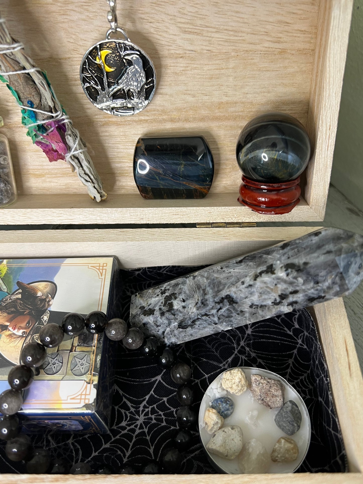 Wooden box filled with crystals, sage, tarot cards, and a crow pendant. 
