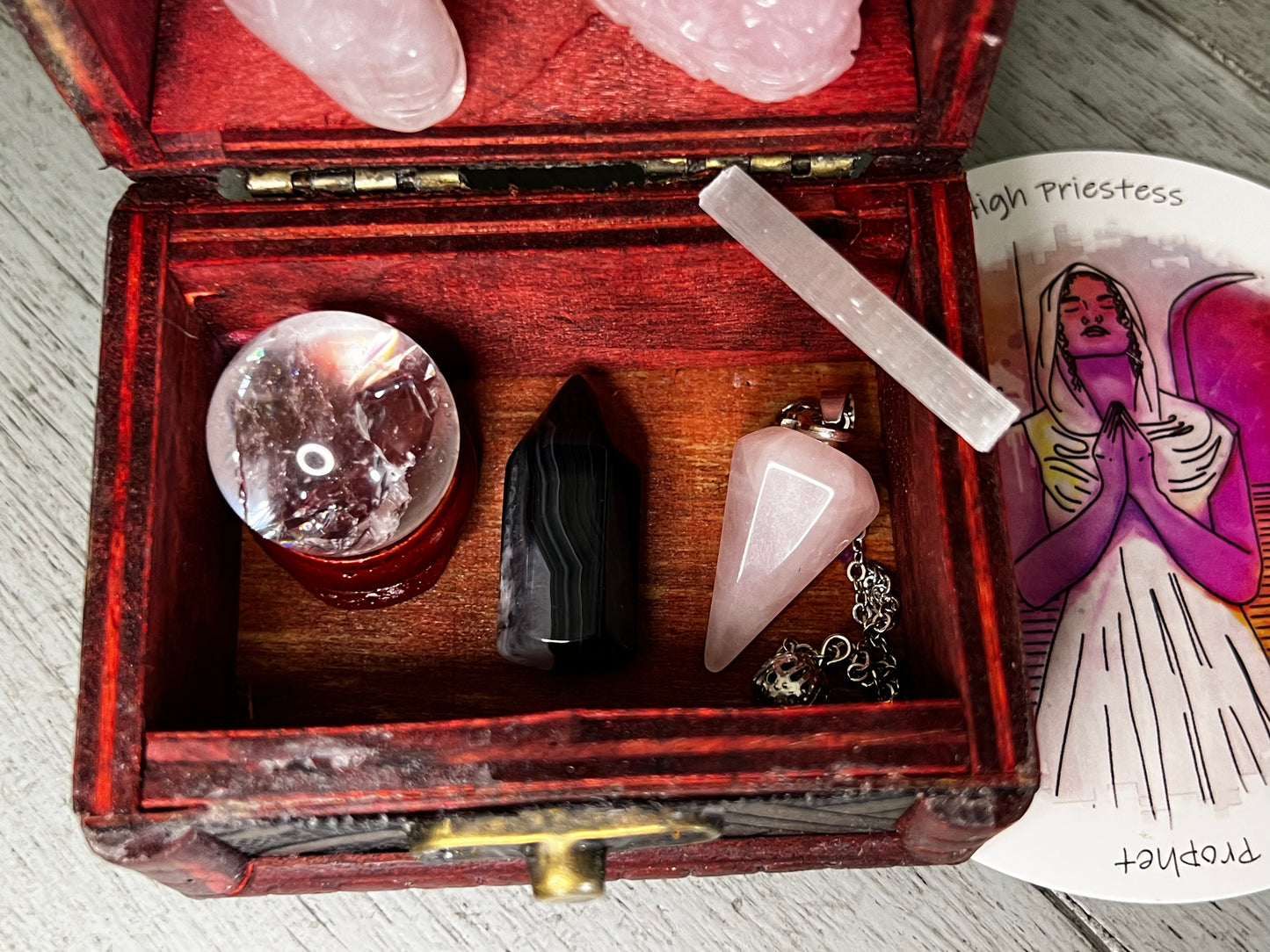 Open red wooden chest containing a crystal ball on a wooden stand, a black and white striped stone, a pink crystal pendulum with a chain, and a white selenite crystal, next to a "High Priestess" tarot card.