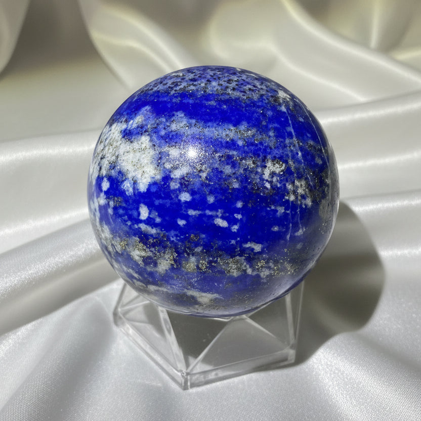 A polished blue lapis lazuli crystal sphere with white and gray veining, displayed on a clear geometric stand 