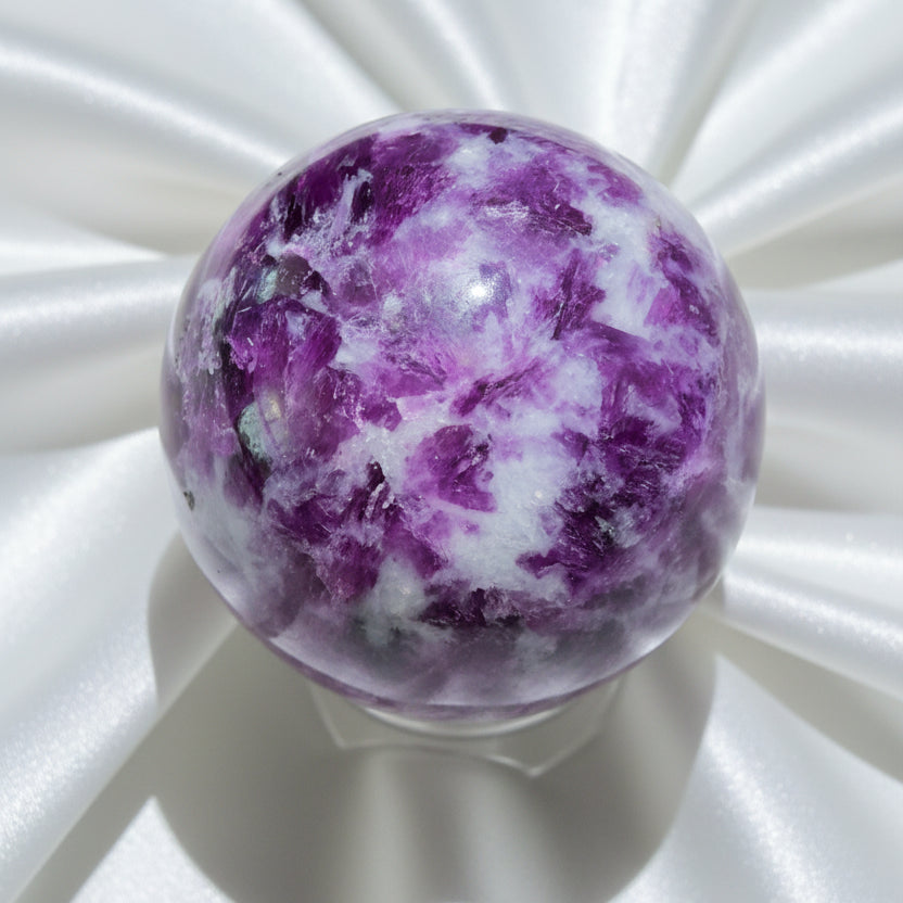 A polished lepidolite purple and white crystal sphere displayed on a clear stand against a light background.