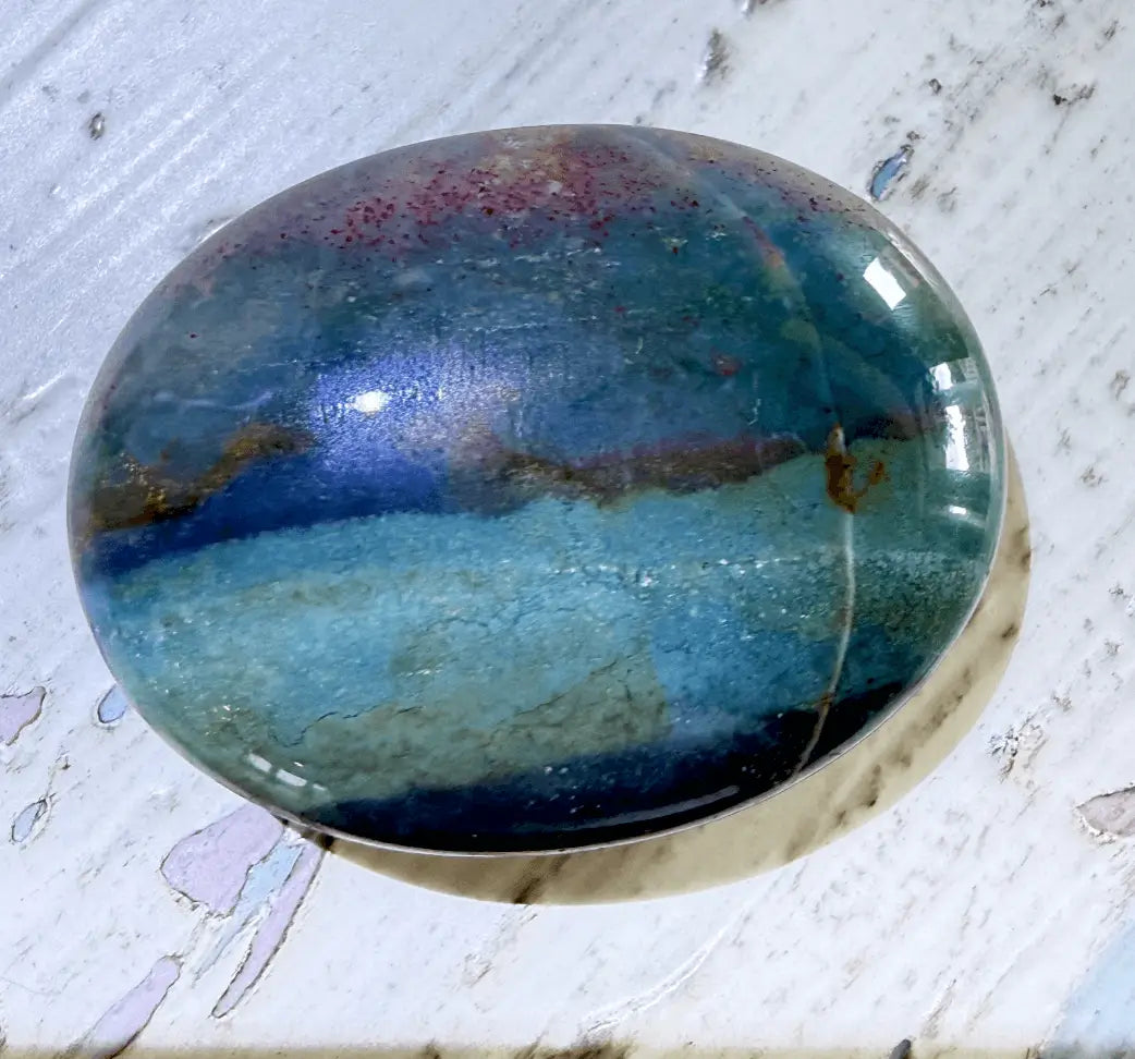 A polished oval ocean jasper cabochon gemstone featuring a marbled pattern of blue, teal, and purple with subtle iridescent shifts, resting on a distressed white wooden surface.