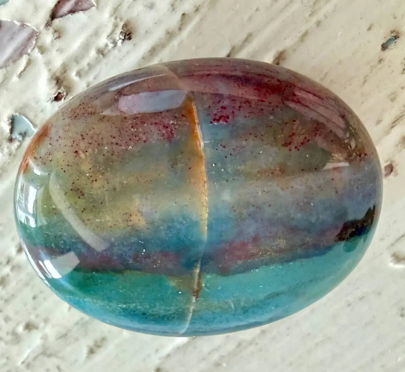 A polished oval ocean jasper palm stone with a marbled pattern of teal, blue, green, beige, and reddish-brown tones, resting on a textured off-white surface.