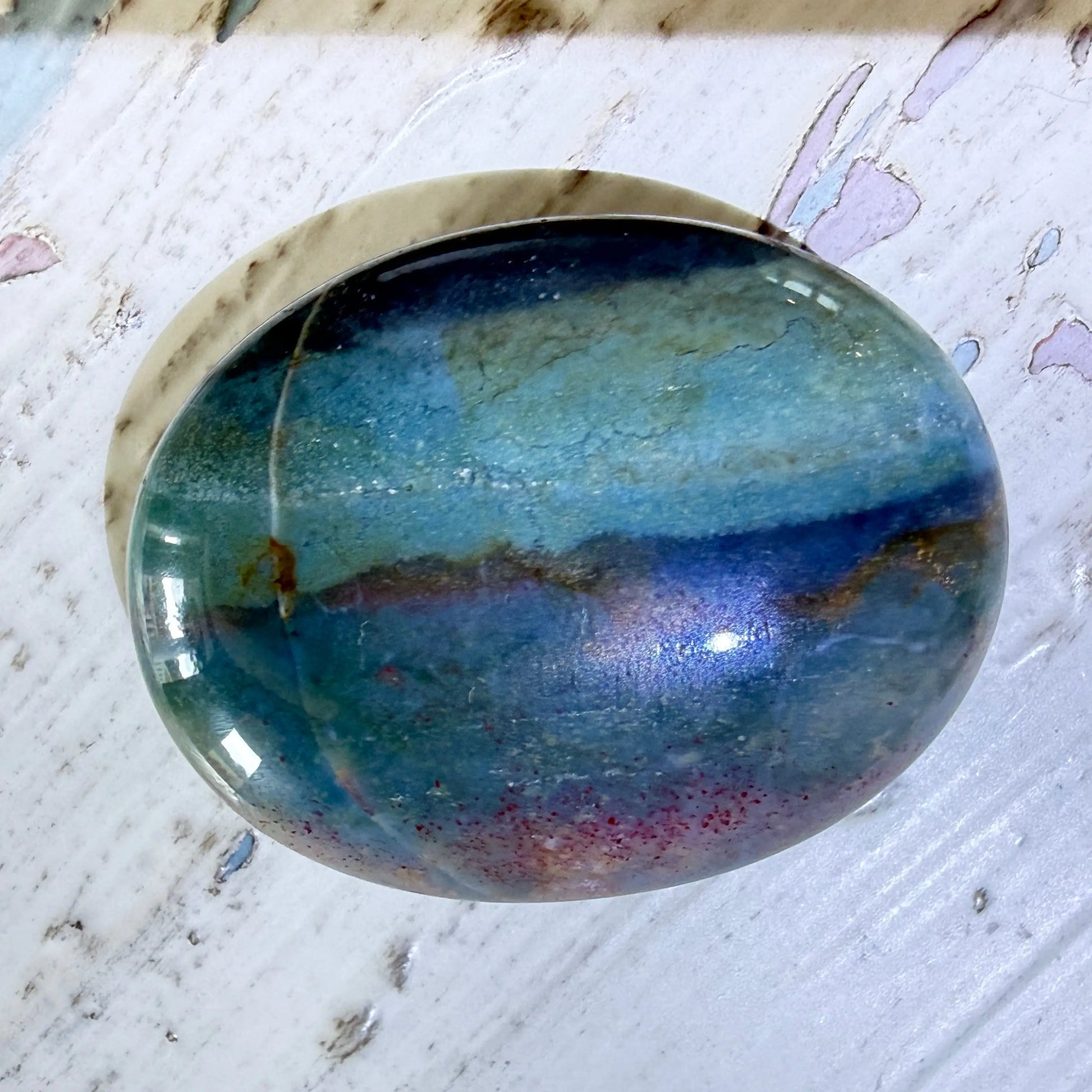 A polished oval ocean jasper palm stone cabochon with a marbled pattern of blue, teal, green, and subtle purple/red veining, resting on a distressed white wooden surface.
