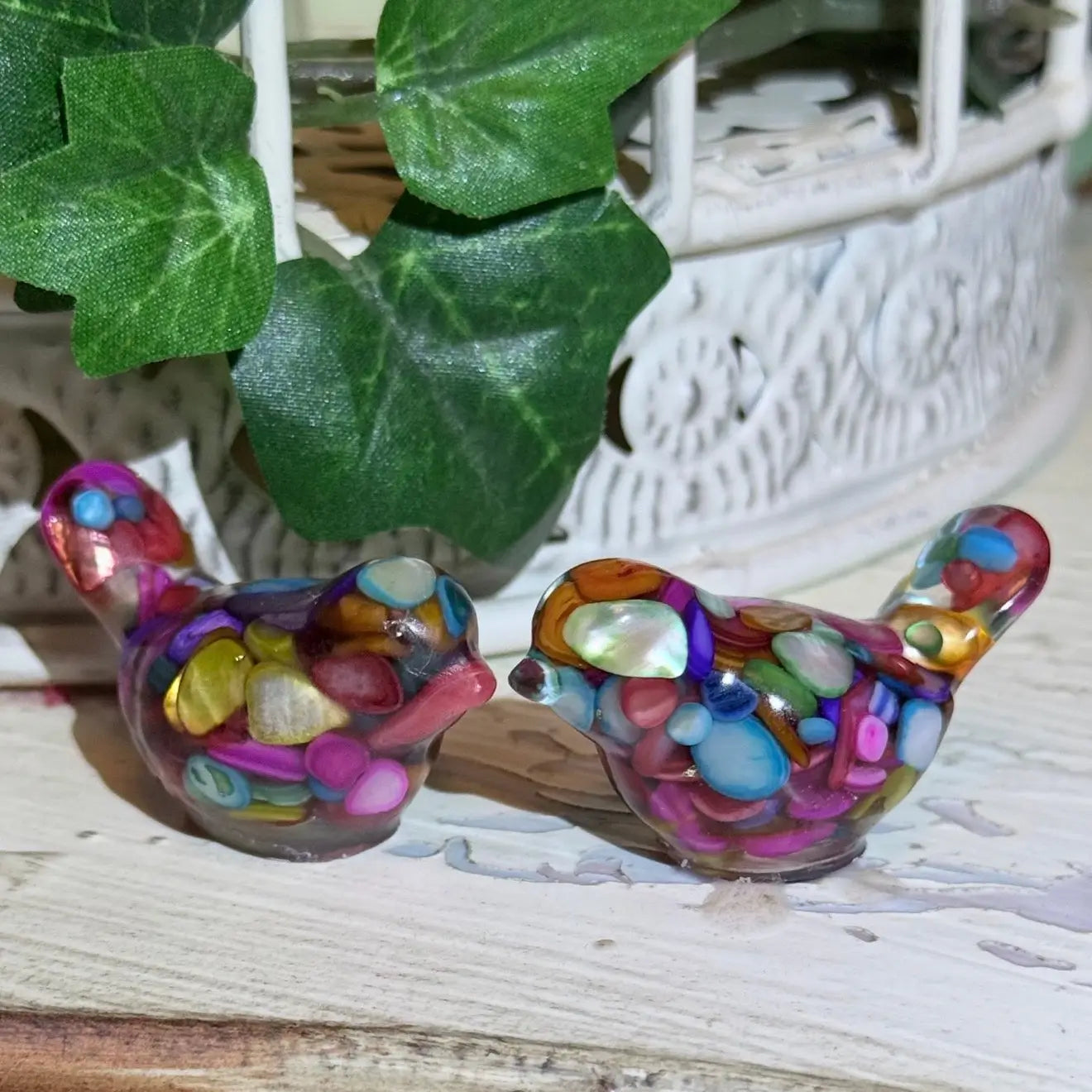 colorful set of two organite-like resin filled birds brightly colored dyed agate gravel and gold tone metal shavings sealed within the resin, sitting on a table with a green ivy beind 