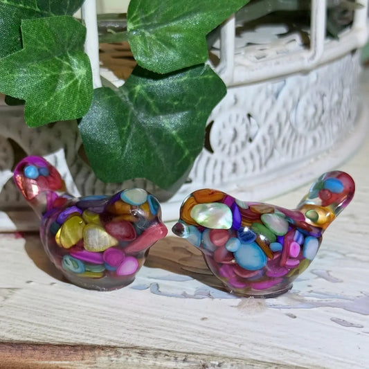 colorful set of two organite-like resin filled birds brightly colored dyed agate gravel and gold tone metal shavings sealed within the resin, sitting on a table with a green ivy beind 