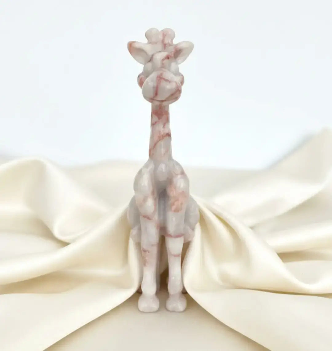 A small sculpted giraffe figurine in pale pink and white marbled stone, standing upright on a cream satin fabric backdrop against a light gray background.