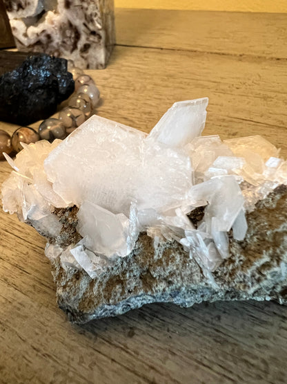 A cluster of white calcite crystals on a rough gray rock surface atop a wood table.
