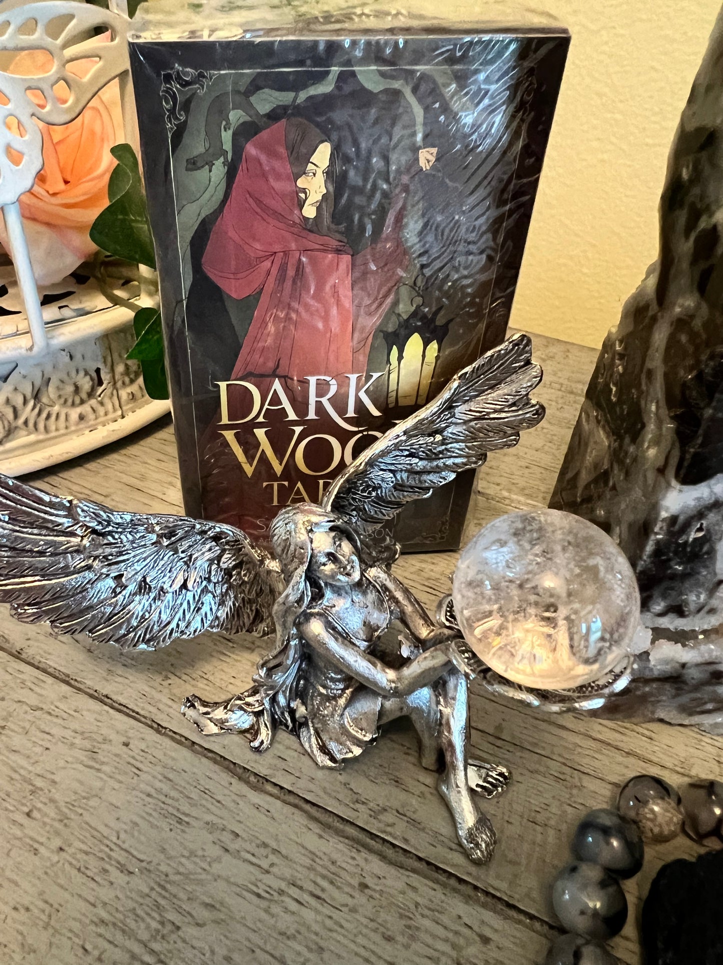 Close-up of a silver fairy statue holding a crystal ball, set against a "Dark Wood Tarot" deck, on a wooden surface.