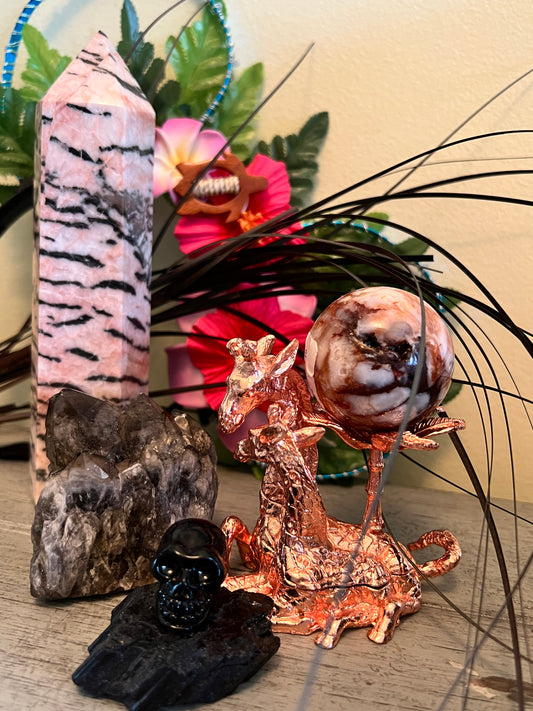 A still life featuring a pink and black striped tower, a cluster of smoky quartz crystals, a black skull figurine on a black rock, a copper giraffe sculpture holding a brown and white marbled orb, and artificial flowers.