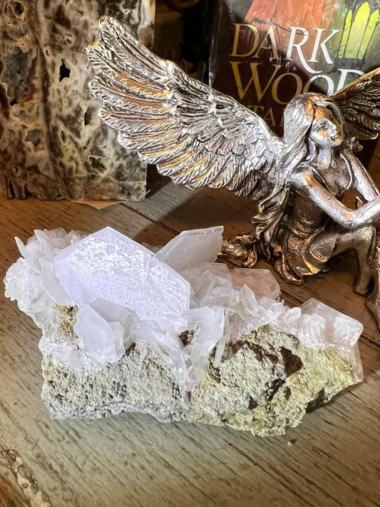 Close-up of a quartz crystal cluster on a rough, earthy matrix, next to a silver angel figurine, on a wooden surface. A book titled "Dark Wood" is in the background.