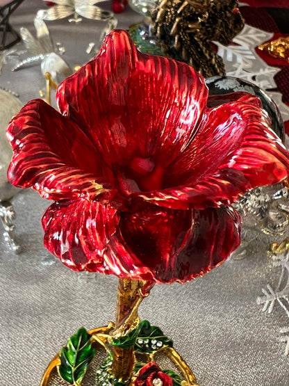 Close-up of a glossy red floral ornament with gold accents, green leaves, and a textured silver background.