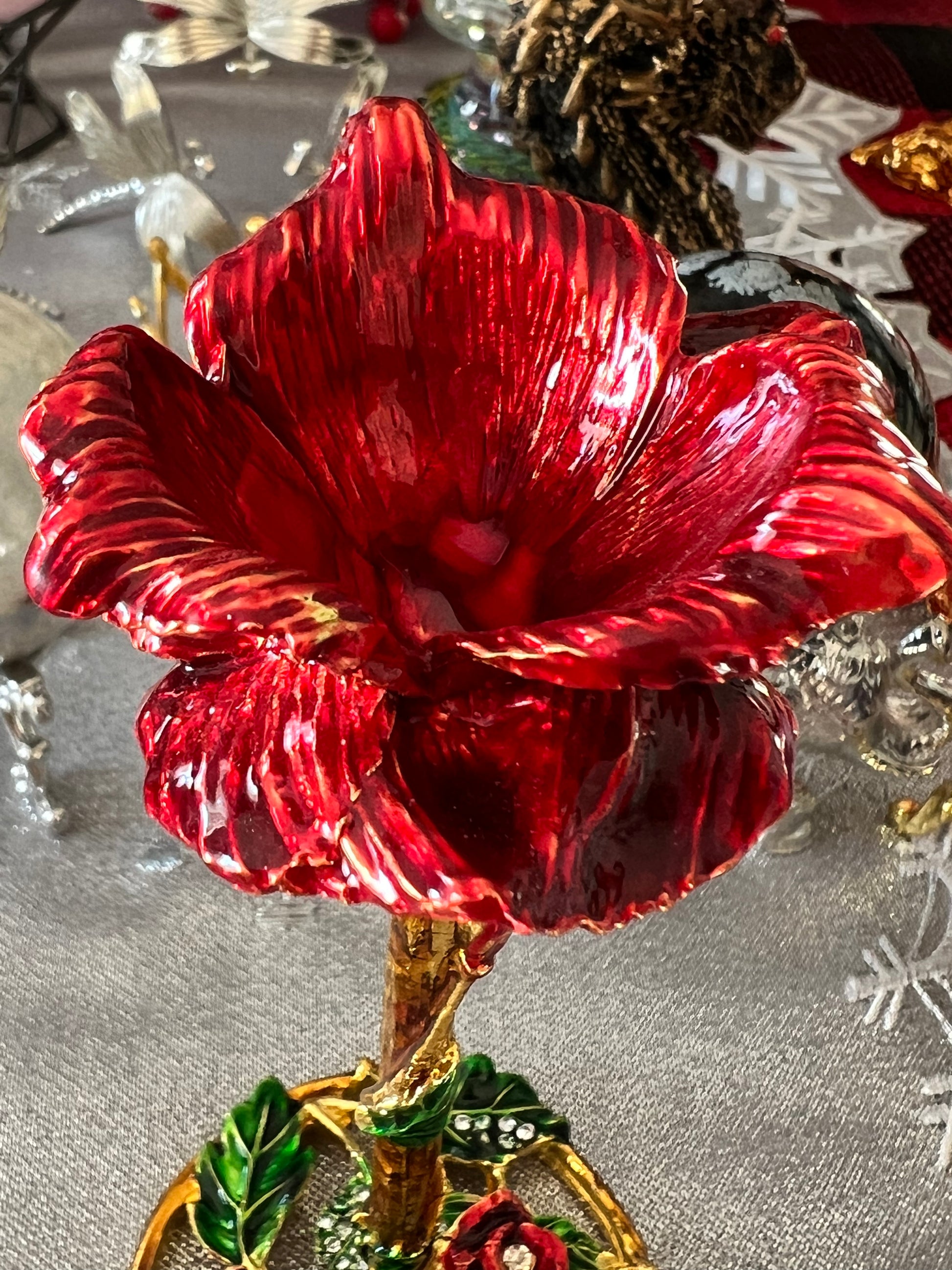 Close-up of a shiny red ornamental flower with a gold stem and green leaves, on a silver tablecloth.
