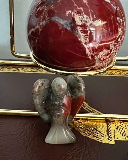 Close-up of a small stone angel figurine and a red, mottled sphere in a gold stand.