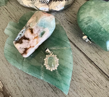 Close-up of various jewelry pieces on a wooden surface. Features a green heart-shaped gemstone, a white and brown layered gemstone, and a silver pendant with a green stone.