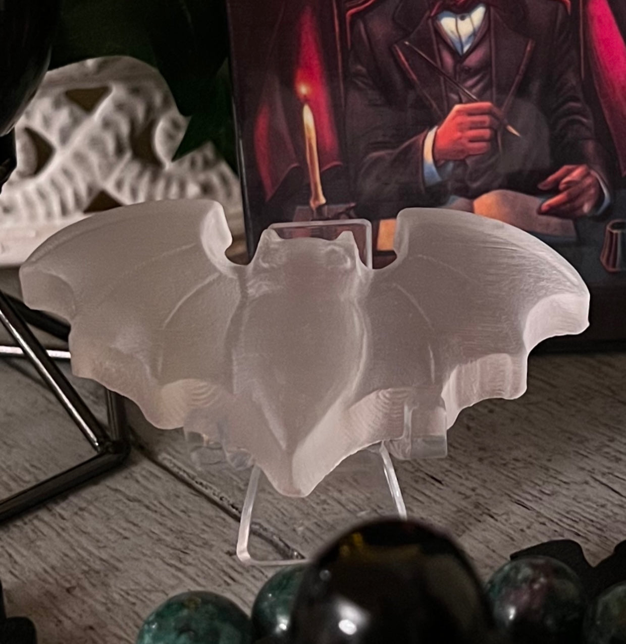 Close-up of a frosted glass bat figurine on a clear stand, set against a dark background with green and black beads in the foreground and a framed picture of a man in a suit in the background.
