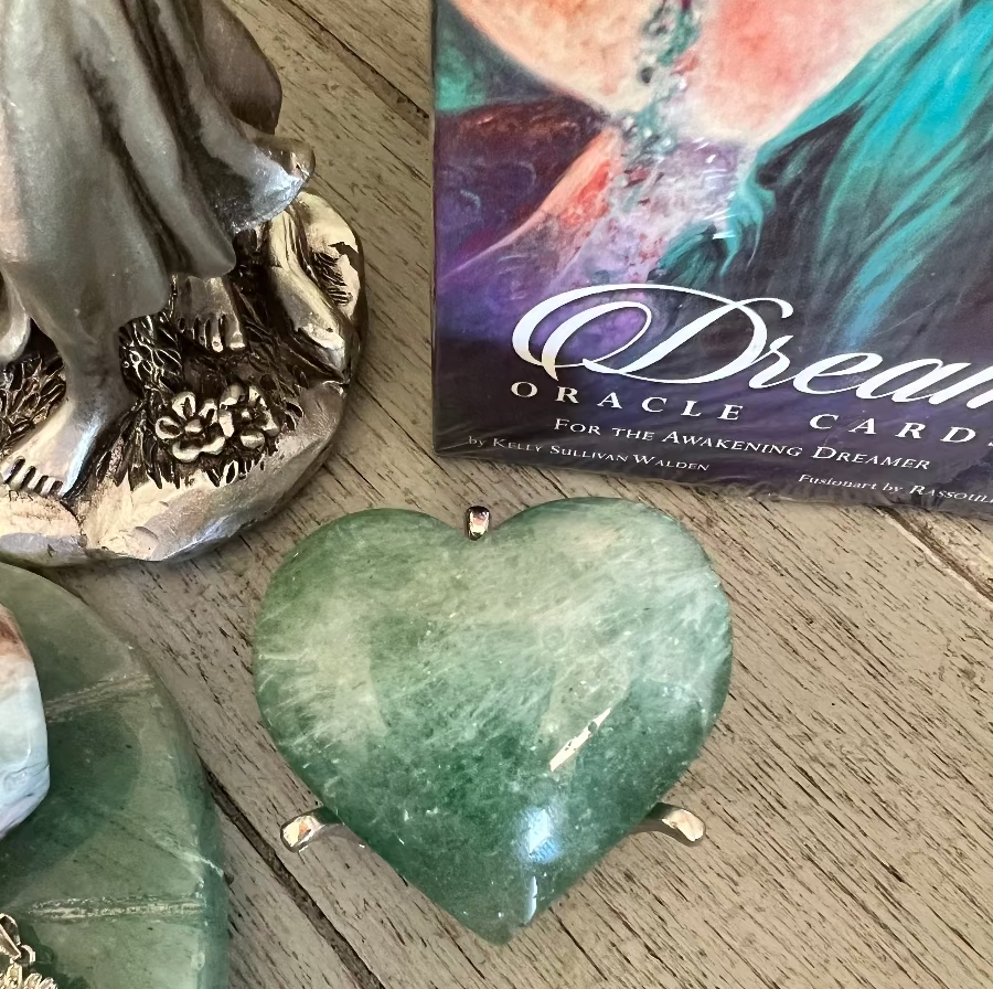 Close-up of a green, heart-shaped crystal, a silver statue base with flowers, and a "Dreams Oracle Cards" box on a weathered wooden surface.
