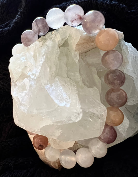 Close-up of a beaded bracelet featuring round, smooth beads in varying shades of white, peach, and purple, resting on a large, jagged chunk of clear quartz against a black fabric background.
