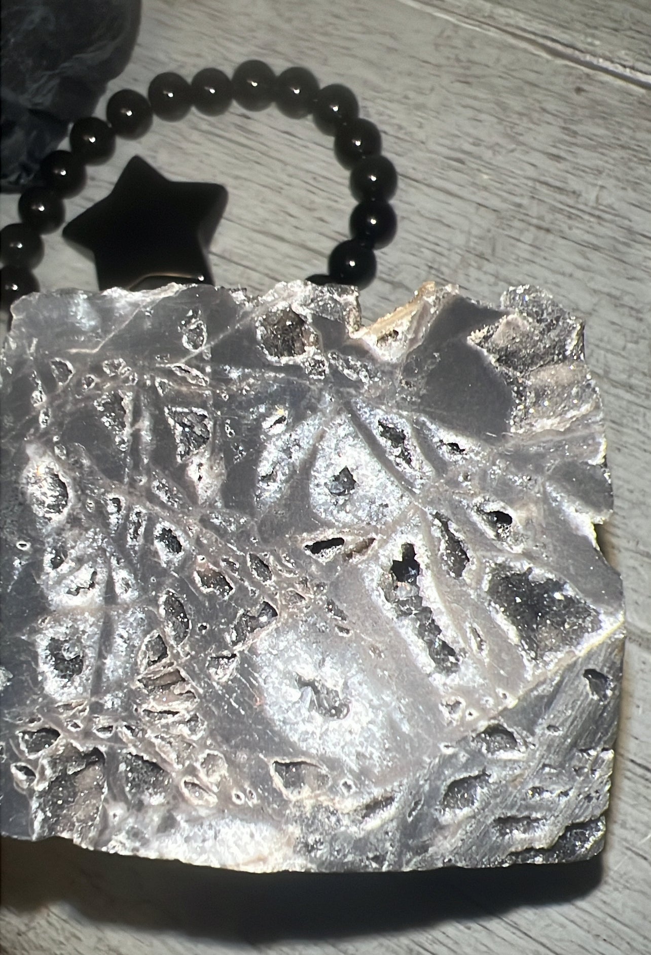 A mineral block with a gray hue, speckled with dark patches, set against a wooden background, with a star pendant and beaded bracelet in the backdrop.