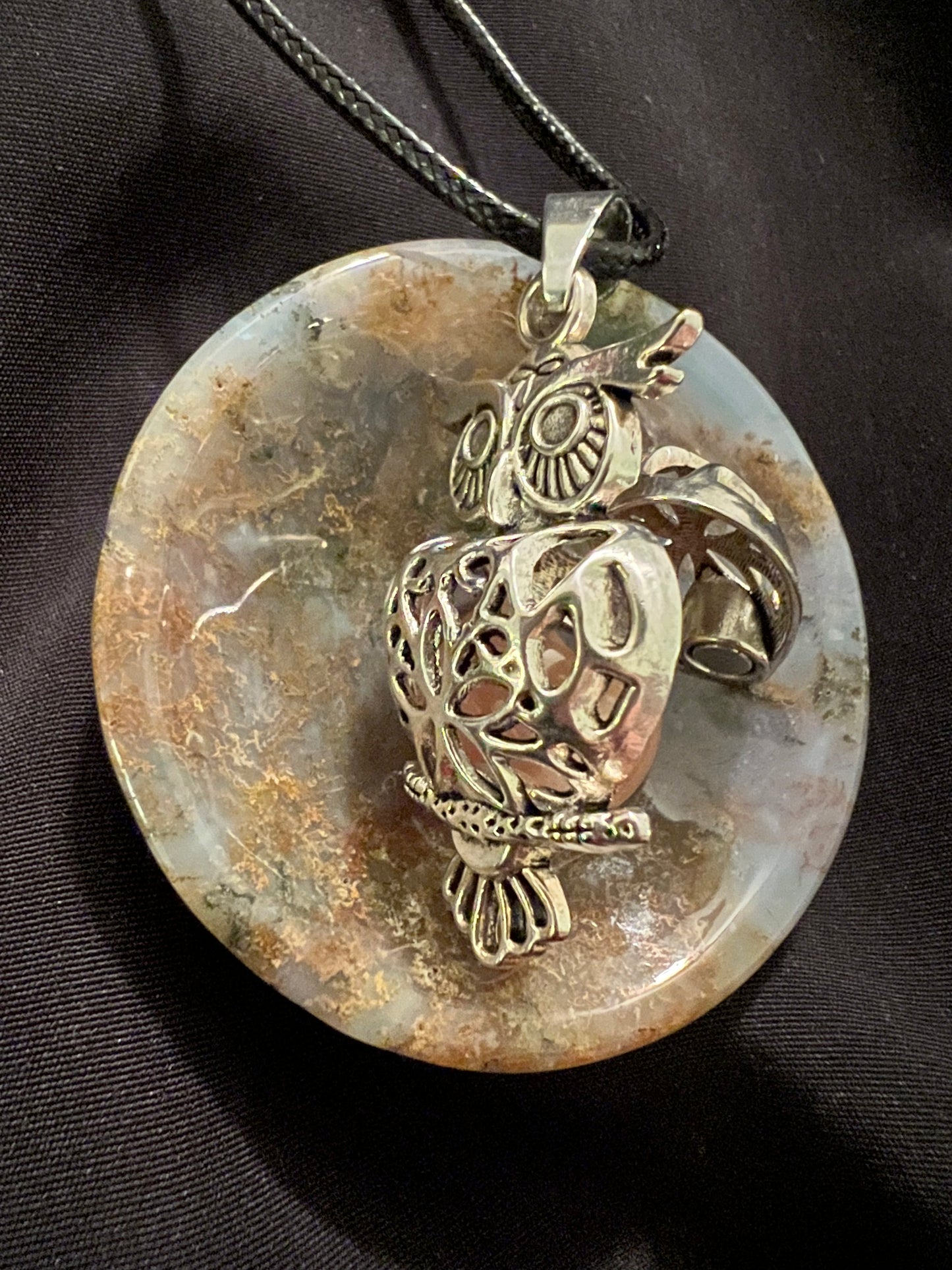 Close-up of an owl pendant necklace featuring silver detailing on a round, polished stone against a black fabric background.