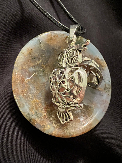 Close-up of an owl pendant necklace featuring silver detailing on a round, polished stone against a black fabric background.