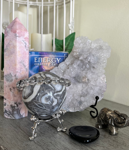 Still life of pink tower crystal, crystal sphere with bracelet, crystal cluster, black polished stone, and elephant figurine on a gray wood surface.